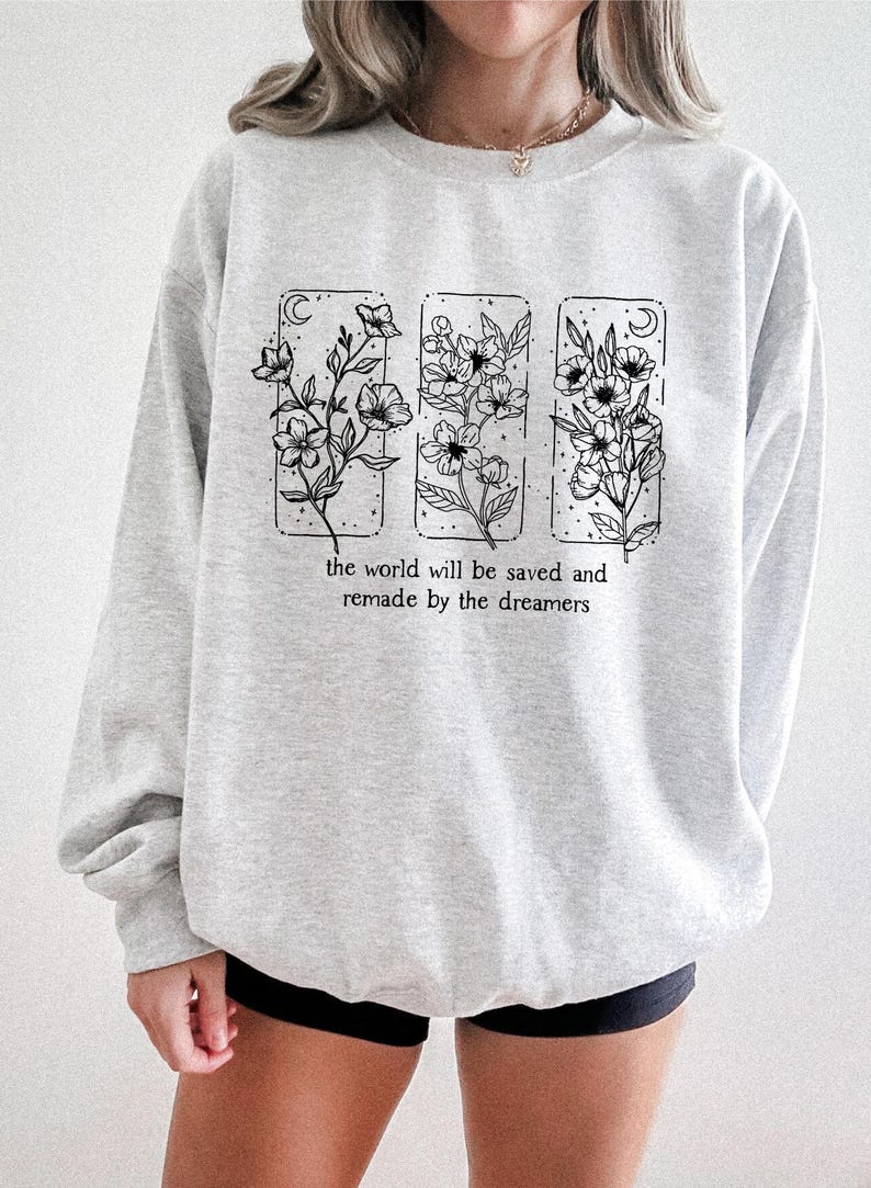 May include: A light gray sweatshirt featuring a floral design in three rectangular frames. Each frame contains a different floral arrangement with a crescent moon. The text "the world will be saved and remade by the dreamers" is printed below the frames.