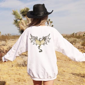 May include: White sweatshirt featuring a detailed dragon graphic on the back. The dragon has outstretched wings with gray and black details, and a yellow and black body. The sweatshirt is worn outdoors in a desert landscape.