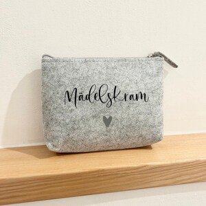 Könnte beinhalten: Eine graue, gepolsterte Kosmetiktasche mit Reißverschluss, auf der "MädelsKram" und ein kleines graues Herz steht.