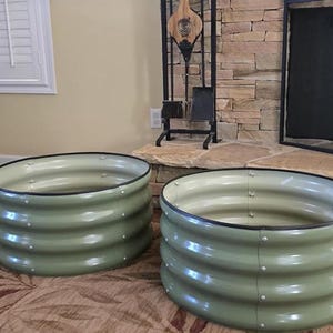 May include: Two round, olive green metal garden planters with a corrugated design. Each planter has a black rim and is secured with silver rivets. The planters are on a patterned rug.