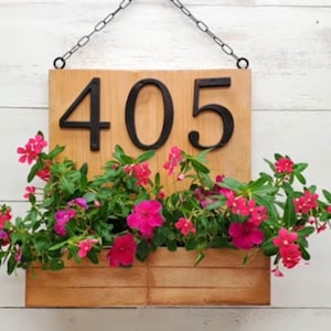 May include: A wooden sign with the number 405 on it, hanging from a chain. A planter box is attached to the bottom of the sign and is filled with pink flowers.