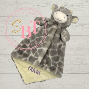 May include: A gray and white giraffe plush security blanket with a yellow fabric underside. The blanket is personalized with the name "Olivia" in pink lettering.