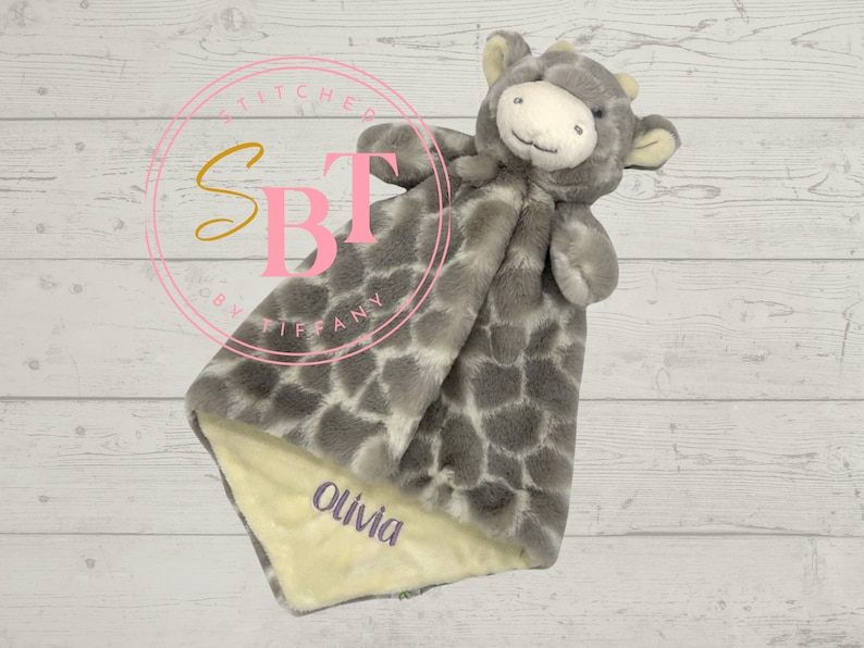 May include: A gray and white giraffe plush security blanket with a yellow fabric underside. The blanket is personalized with the name "Olivia" in pink lettering.