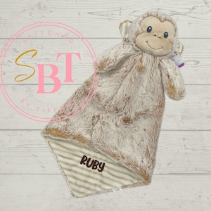 May include: A plush monkey security blanket with a brown and beige fur texture. The blanket has a white and brown striped lining with the name "Ruby" embroidered on it.