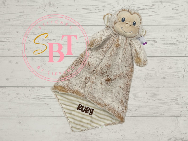 May include: A plush monkey security blanket with a brown and beige fur texture. The blanket has a white and brown striped lining with the name "Ruby" embroidered on it.