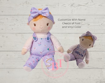 Puede incluir: Dos muñecas de peluche con cabello naranja, vestidas con petos florales color lavanda y diademas a juego. La muñeca más grande está de pie y la más pequeña sentada, con el nombre Olivia en texto azul oscuro. Texto: Customize With Name Choice of Font and Vinyl Color.