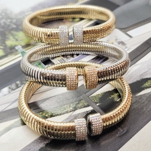 May include: Three gold and silver-toned bracelets. Each bracelet has a ribbed design and a clasp adorned with small, sparkling stones. The bracelets are stacked, showcasing their varying metallic finishes and intricate details.