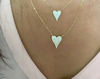 May include: Two gold necklaces with heart-shaped pendants. The pendants are encrusted with small, sparkling stones. The necklaces are layered, with one pendant slightly smaller than the other. The chains are delicate and the hearts are pointed at the bottom.