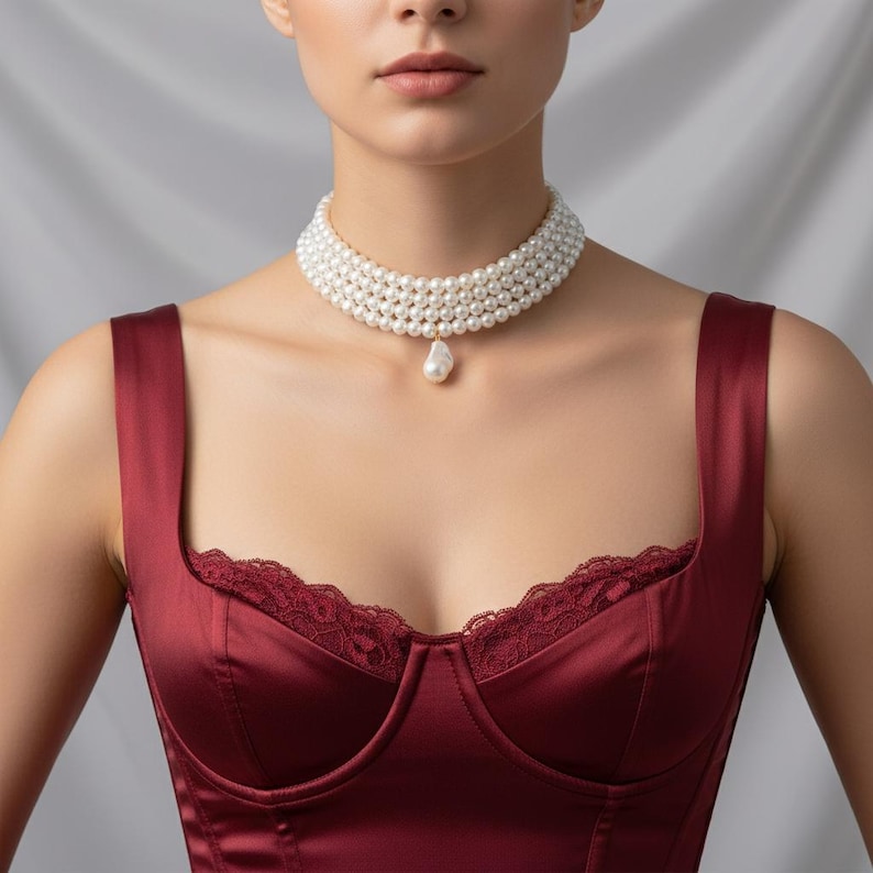 May include: A multi-strand pearl choker necklace with a single, teardrop-shaped pearl pendant. The necklace is worn over a burgundy satin top with lace trim. The pearls are white and the top is a deep red color.