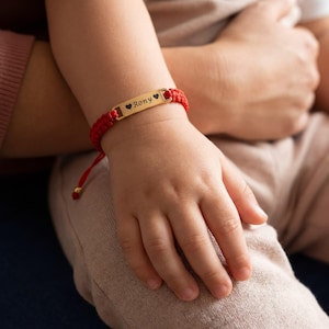 Puede incluir: Una pulsera trenzada roja con una placa rectangular dorada. La placa está grabada con el nombre "Rony" y dos símbolos de corazón. La pulsera está en la muñeca de un niño.
