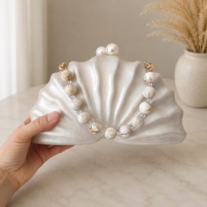 May include: A shell-shaped clutch bag with a glossy off-white finish and a ridged surface. The bag features a top clasp with two large white pearl-like spheres and a decorative handle made of marbled beige and white spherical beads on gold links.