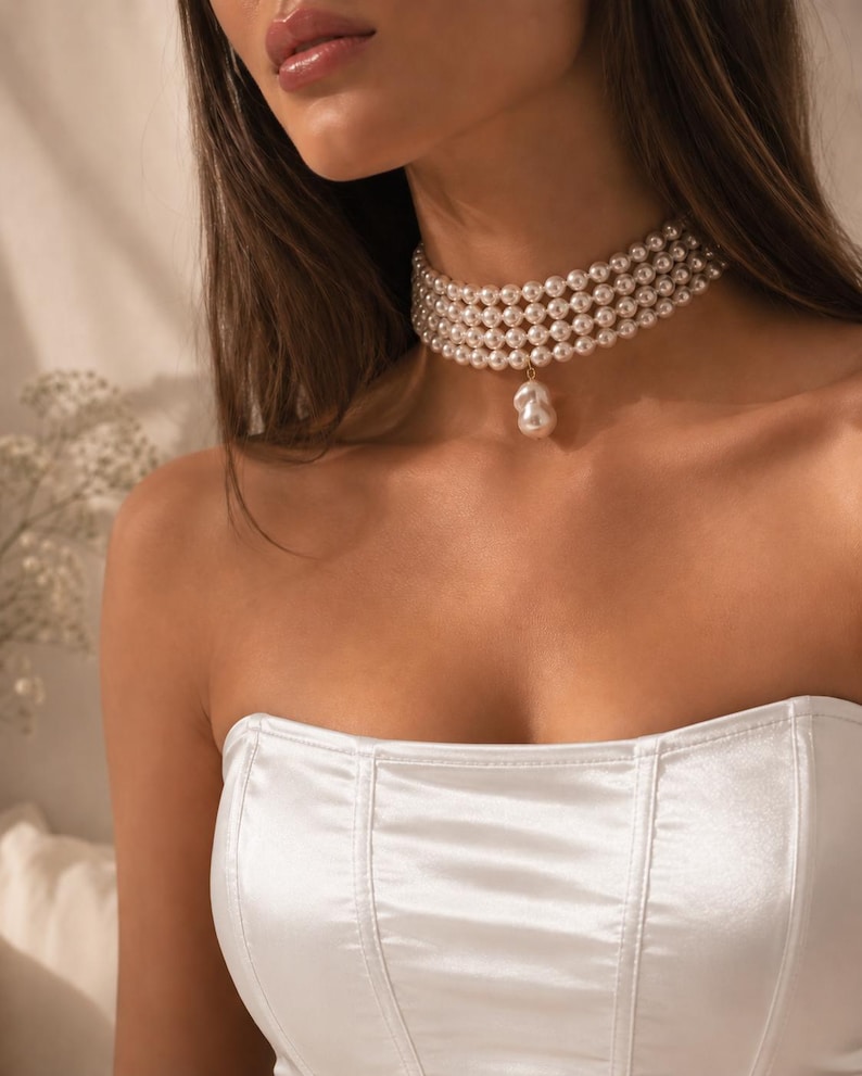 May include: A multi-strand pearl choker necklace with a large, single pearl pendant. The necklace is composed of several rows of small, round pearls. The model is wearing a white strapless top.