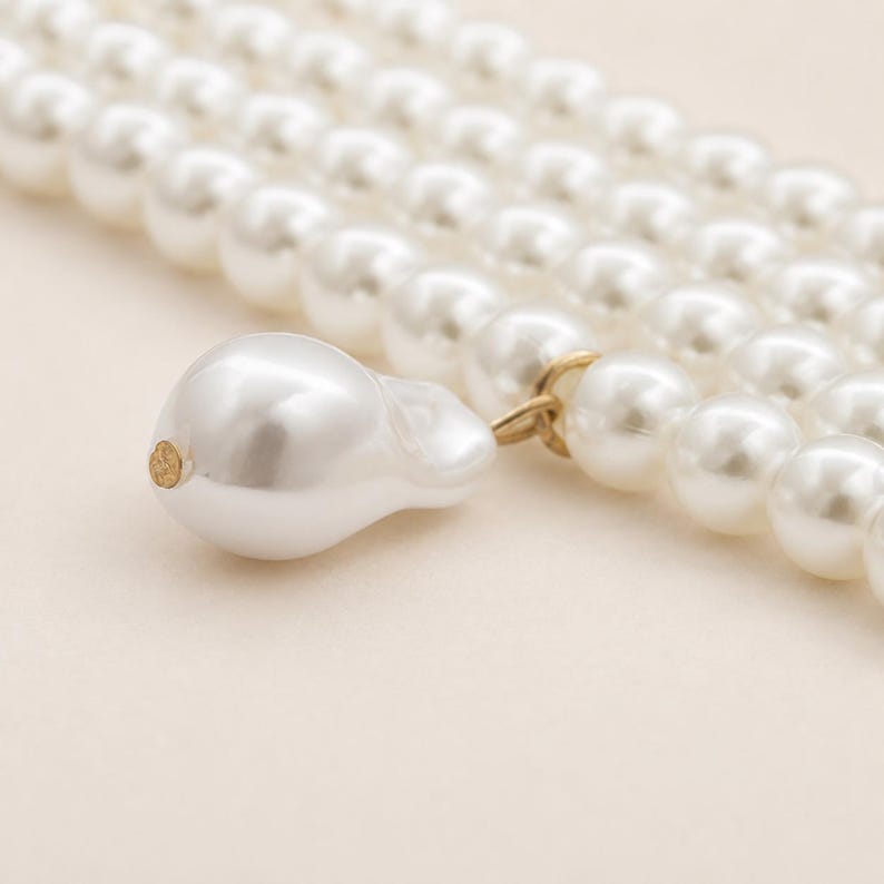 May include: Close-up of a white pearl necklace with a large, irregular pearl pendant. The necklace features multiple strands of round, lustrous pearls. The pendant is connected with a gold-colored clasp, adding a touch of elegance to the jewelry.