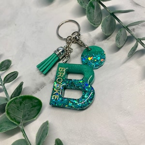 Op de afbeelding: Een groene glitter sleutelhanger met een zilveren bedel en een kwast. De sleutelhanger is gevormd als de letter "B" en heeft de naam "Brooke" erop.