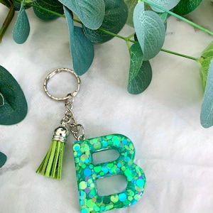 Puede incluir: Un llavero de purpurina verde y azul con la letra "B" y un borla verde.