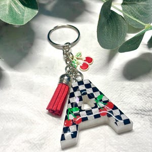 Puede incluir: Un llavero decorativo con la letra "A" en un patrón de cuadros blanco y negro, adornado con cerezas rojas. Incluye una borla roja y un llavero plateado. Un accesorio personalizado.