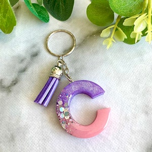 Puede incluir: Un llavero de resina con forma de letra C, de color morado y rosa, con purpurina plateada y un borla morado.