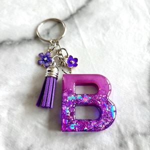 May include: A purple resin keychain in the shape of the letter "B". The keychain has a silver ring, a purple tassel, and purple flower charms. The letter is filled with iridescent glitter.