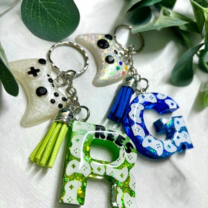 May include: Three white and green resin keychains with a gamer controller design. The keychains spell out the word "REES" with a blue keychain with a gamer controller design and the letter "G" hanging from the top.