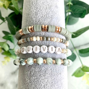May include: A set of four bracelets featuring a green and white marble pattern, a white bead bracelet with the name "OLIVIA" spelled out, a gray beaded bracelet, and a gold chain bracelet.