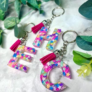 Può includere: Tre ciondoli portachiavi in resina a forma delle lettere E, H e C. Ogni lettera è riempita di coriandoli e glitter colorati e ha un fiocco rosa attaccato alla parte superiore.