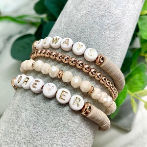 Op de afbeelding: Een set van vijf armbanden met witte kralen en gouden accenten. De armbanden spellen de woorden "FOREVER" en "ALWAYS" uit.