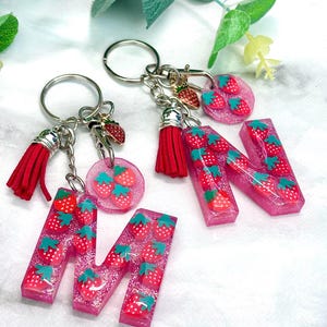 May include: Pink glitter resin keychains featuring the letters "M" and "N", each filled with strawberry charms. The keychains have red tassels and silver key rings. A pink circular charm with strawberries is also included.