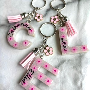 Può includere: Quattro portachiavi con lettere personalizzate. Ogni lettera è decorata con fiori rosa e un nome in corsivo. I portachiavi hanno un nappa rosa e un ciondolo a forma di fiore. Le lettere sono "C", "L" e "E".