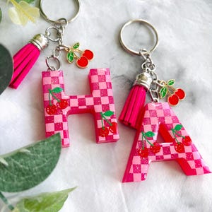 Puede incluir: Dos llaveros de resina rosa, uno con forma de letra "H" y otro "A". Ambos tienen un patrón de cuadros y detalles de cerezas. Cada llavero tiene una borla rosa y un llavero plateado.