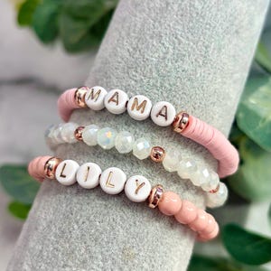 Op de afbeelding: Twee roze kralenarmbanden met witte kralen die de namen "MAMA" en "LILY" in gouden letters spellen.
