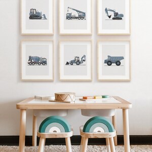 Op de afbeelding: Zes ingelijste prints van verschillende bouwvoertuigen in tinten blauw en grijs, hangend aan een witte muur boven een houten tafel met twee regenboogstoelen.
