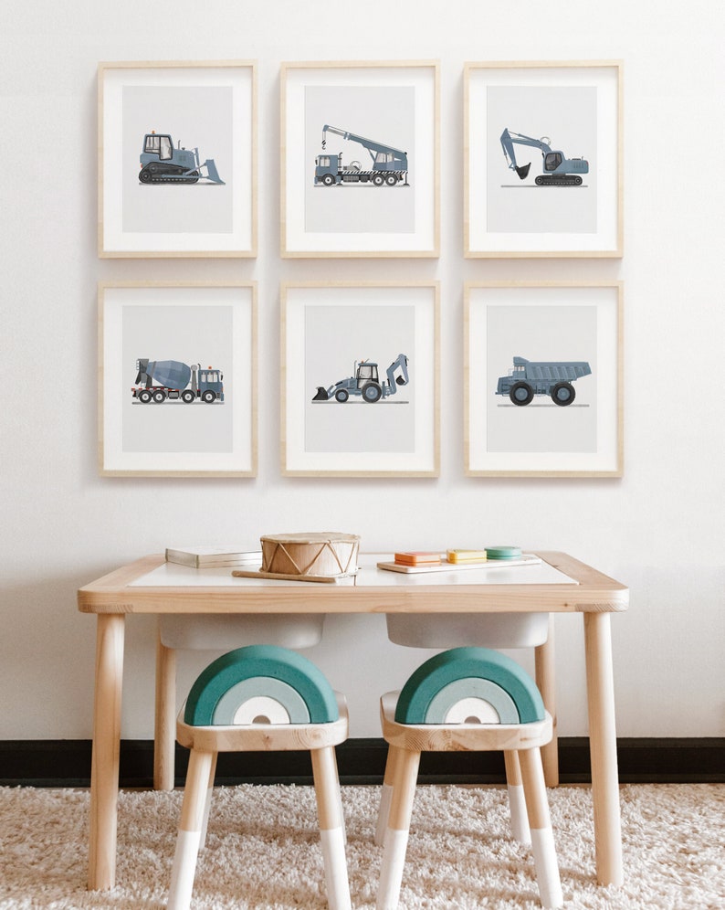 Op de afbeelding: Zes ingelijste prints van verschillende bouwvoertuigen in tinten blauw en grijs, hangend aan een witte muur boven een houten tafel met twee regenboogstoelen.