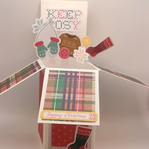 May include: A pop-up card with a plaid pattern and the text "Happy Christmas". The card features a variety of winter-themed decorations, including a snowflake, a mitten, a sweater, and a stocking.