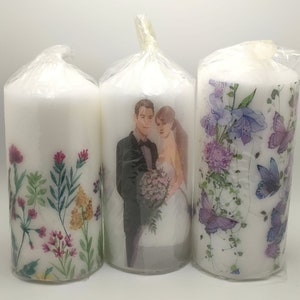 May include: Three white pillar candles with different designs. The first candle features a floral design with pink, yellow, and green flowers. The second candle has a photograph of a couple in wedding attire. The third candle features a floral design with purple flowers and butterflies.