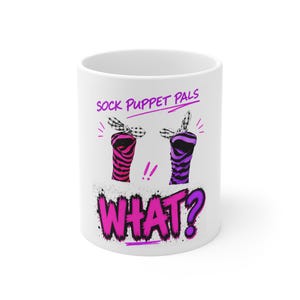 May include: A white ceramic mug featuring a graphic of two sock puppets with black and white checkered bows. One puppet is pink and black striped, the other is purple and black striped. The text reads SOCK PUPPET PALS and WHAT? in pink.