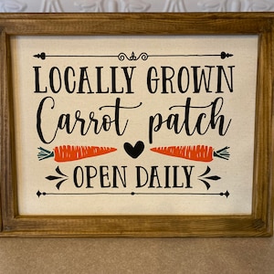 May include: A wooden framed sign with a white canvas background. The sign reads "Locally Grown Carrot Patch Open Daily" with two orange carrots and a black heart in the middle.