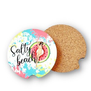 May include: A round, cork-backed coaster with a colorful design featuring a woman in a swimsuit floating on a watermelon. The text "Salty Beach" is written in black on the coaster.
