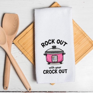 May include: White kitchen towel with the text "ROCK OUT with your CROCK OUT" and a pink slow cooker graphic. The towel is on a wooden cutting board with two wooden spoons.