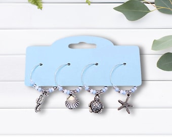 May include: Set of four glass charms with silver seahorse, seashell, turtle, and starfish charms. The charms are attached to silver rings with blue and white beads. The charms are displayed on a light blue card with a hole at the top.