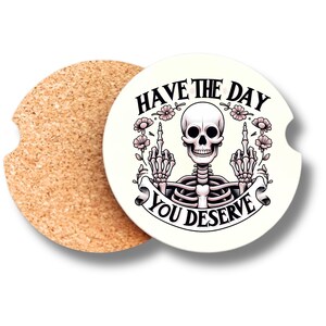 May include: A round car coaster with a skeleton graphic giving the middle finger, surrounded by flowers. The text reads "HAVE THE DAY YOU DESERVE". A second, plain cork coaster is next to it.