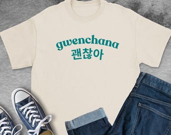 May include: A cream-colored short-sleeve t-shirt featuring the text "gwenchana" in a dark teal arched font, with the Korean characters "괜찮아" printed directly underneath in the same dark teal color and font style.