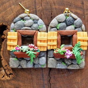 May include: A pair of whimsical earrings featuring miniature stone houses with brown window frames and yellow shutters. Each house has a flower box with a frog, mushrooms, and flowers. The earrings hang from gold-colored hooks.