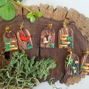 May include: Handmade polymer clay earrings featuring a wood grain design with gold-colored ear wires. Each earring showcases a different woodland creature or element, including a red cardinal, squirrel, mushrooms, and a green lizard.