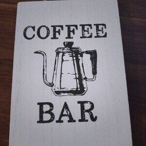 May include: A rectangular white wooden sign with a jute twine hanger. The sign features the black text COFFEE BAR and a black graphic illustration of a gooseneck kettle centered between the words. The wood grain is visible through the white paint.