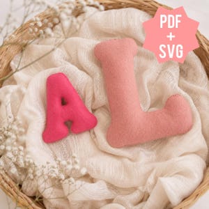 May include: Two plush felt letters, a hot pink A and a light pink L, resting on white fabric. A pink star-shaped graphic in the upper right corner contains the white text PDF + SVG. The letters have a soft, rounded, and padded appearance.