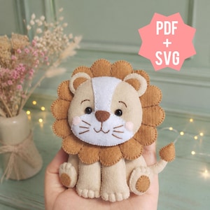 Può includere: Un peluche a forma di leone in feltro con corpo color marrone chiaro, criniera smerlata marrone, macchia bianca sul viso e naso marrone. Il giocattolo ha occhi a perla neri, baffi ricamati e guance rosa. Un grafico rosa indica PDF + SVG.