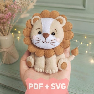 Könnte beinhalten: Ein Filz-Löwenornament mit beigem Körper, brauner, gewellter Mähne und weißem Gesichtsteil mit schwarzen, gestickten Augen, Nase und Schnurrhaaren. Der Löwe hat einen kleinen Schwanz und rosa Wangen. Ein rosa Banner unten zeigt PDF + SVG.