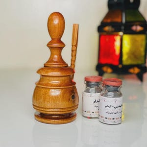 Puede incluir: Un quemador de incienso de madera con un diseño decorativo, acompañado de una herramienta de madera y dos pequeños frascos de vidrio con tapas rojas. Los frascos tienen etiquetas con texto. Una linterna colorida en el fondo.