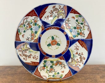 May include: A large, round, white ceramic plate with a blue and red geometric pattern and floral designs. The plate has a raised rim and is decorated with a variety of flowers, leaves, and branches. The plate is on a wooden surface.