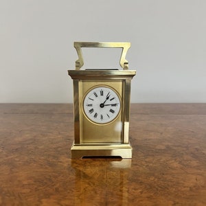 Può includere: Un piccolo orologio rettangolare in ottone con un quadrante bianco e indici orari romani neri. L'orologio ha una maniglia nella parte superiore e si trova su una superficie in legno.